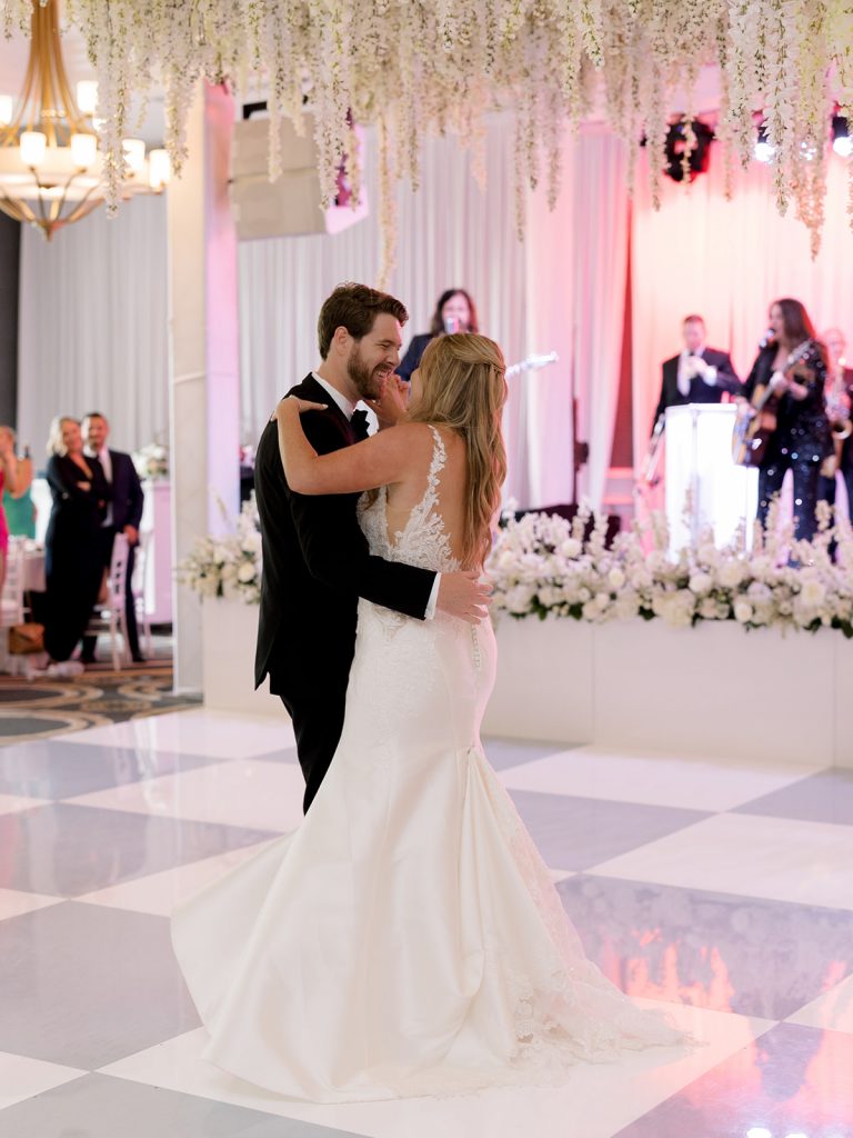 A classic meets modern, white wedding at the Austin Country Club. This wedding reception features luxe white wisteria dripping from the ceiling, a garden style design aesthetic, lots of candles, Stage floral, and a head table overhead installation flanking the dance floor. the bride and groom are danging In Austin Texas