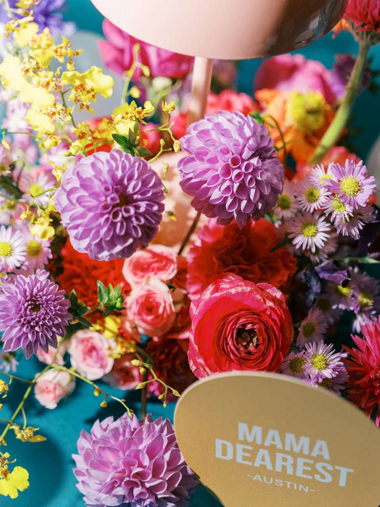 colorful wedding floral centeprieces on a dining table with a table lamp and bar sign table numbers. Flowers like Dahlias, ranunculus, and orchids are seen in this tablescape.\